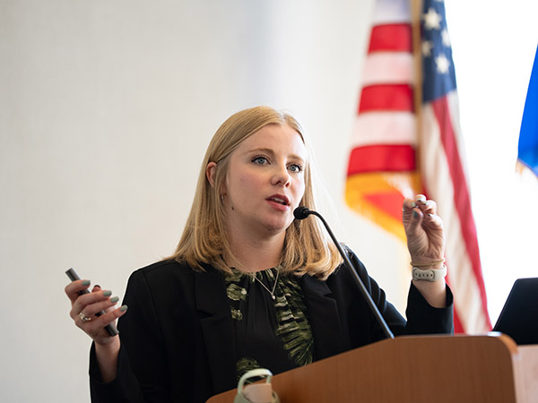 photo of woman speaking at lectern, flags in background