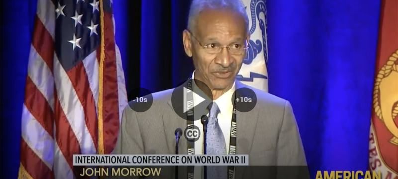photo of man speaking at microphone, with flags and lower thirds text