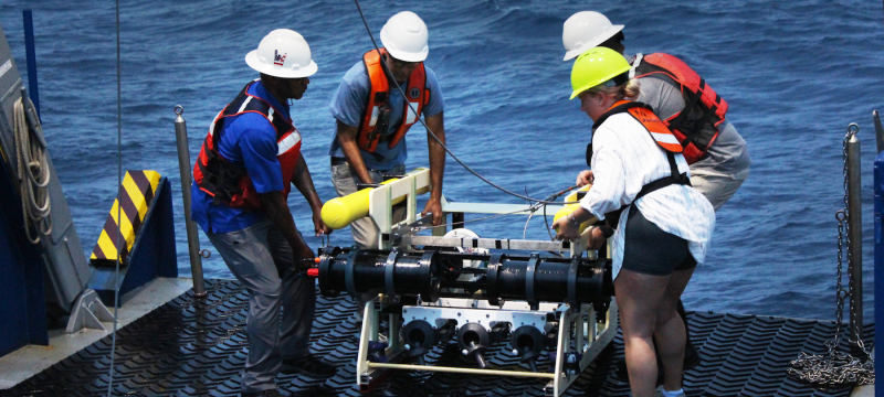 photo off four people with large device in deck of ship, day, water in background