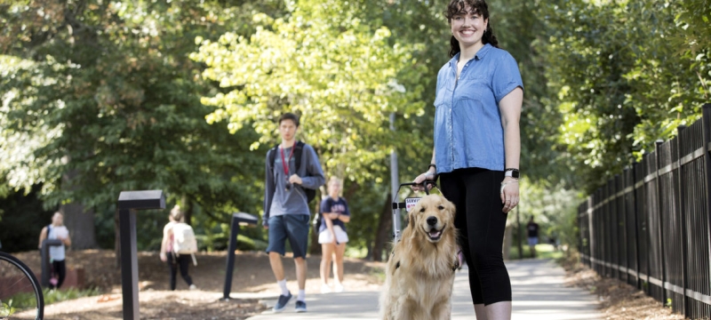 outdoor photo of woman with service dog on sidewalk and other pedestrians.